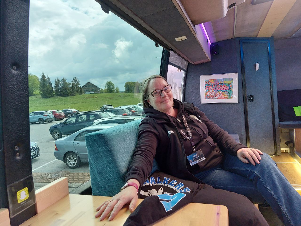 Hymyilevä nainen istuu bussissa, taustalla parkkeerattuja autoja ja vihreä pelto