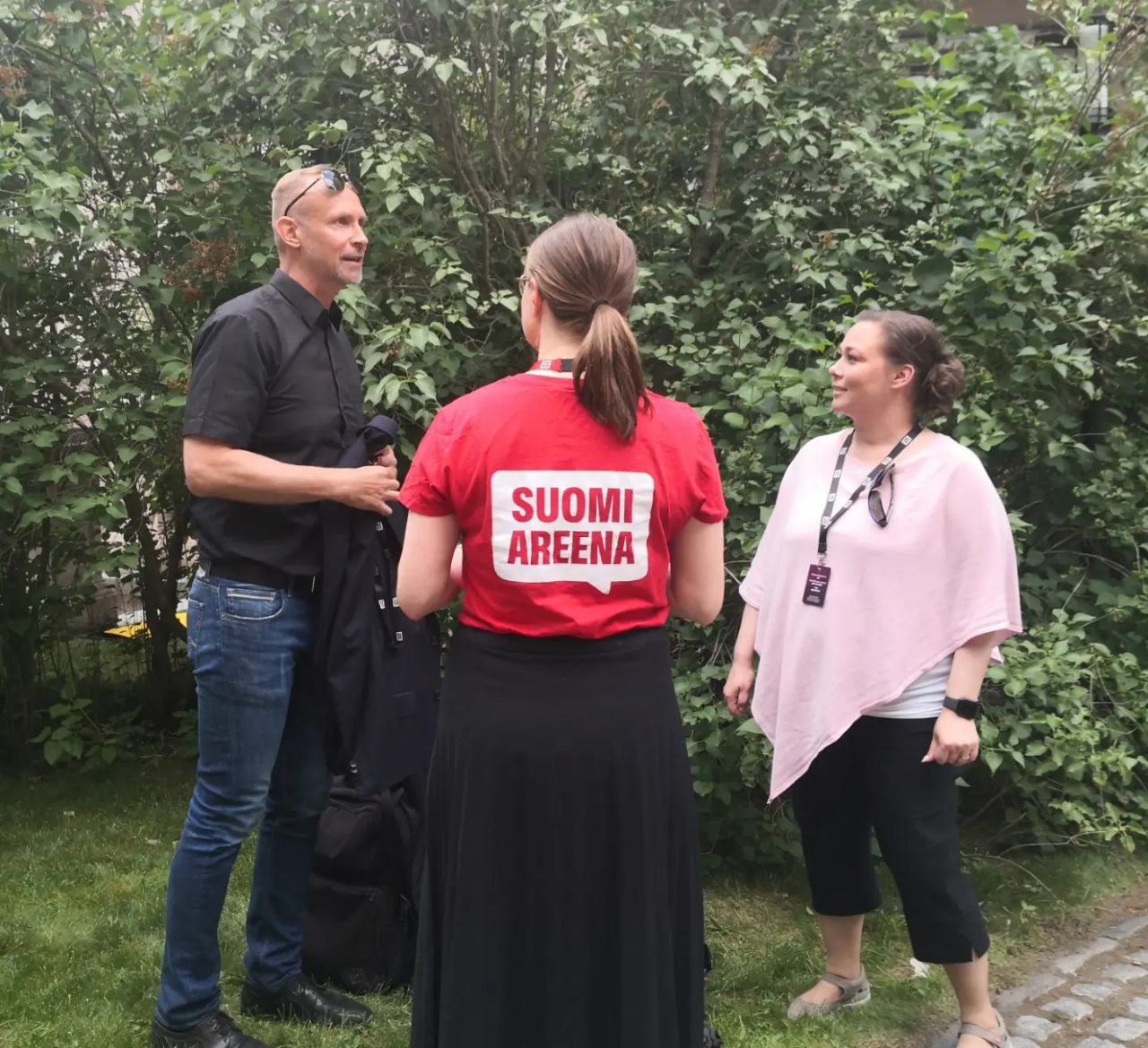 Kolme ihmistä keskustelee vihreässä ympäristössä, keskimmäinen selin kameraan punaisessa SuomiAreena-paidassa.