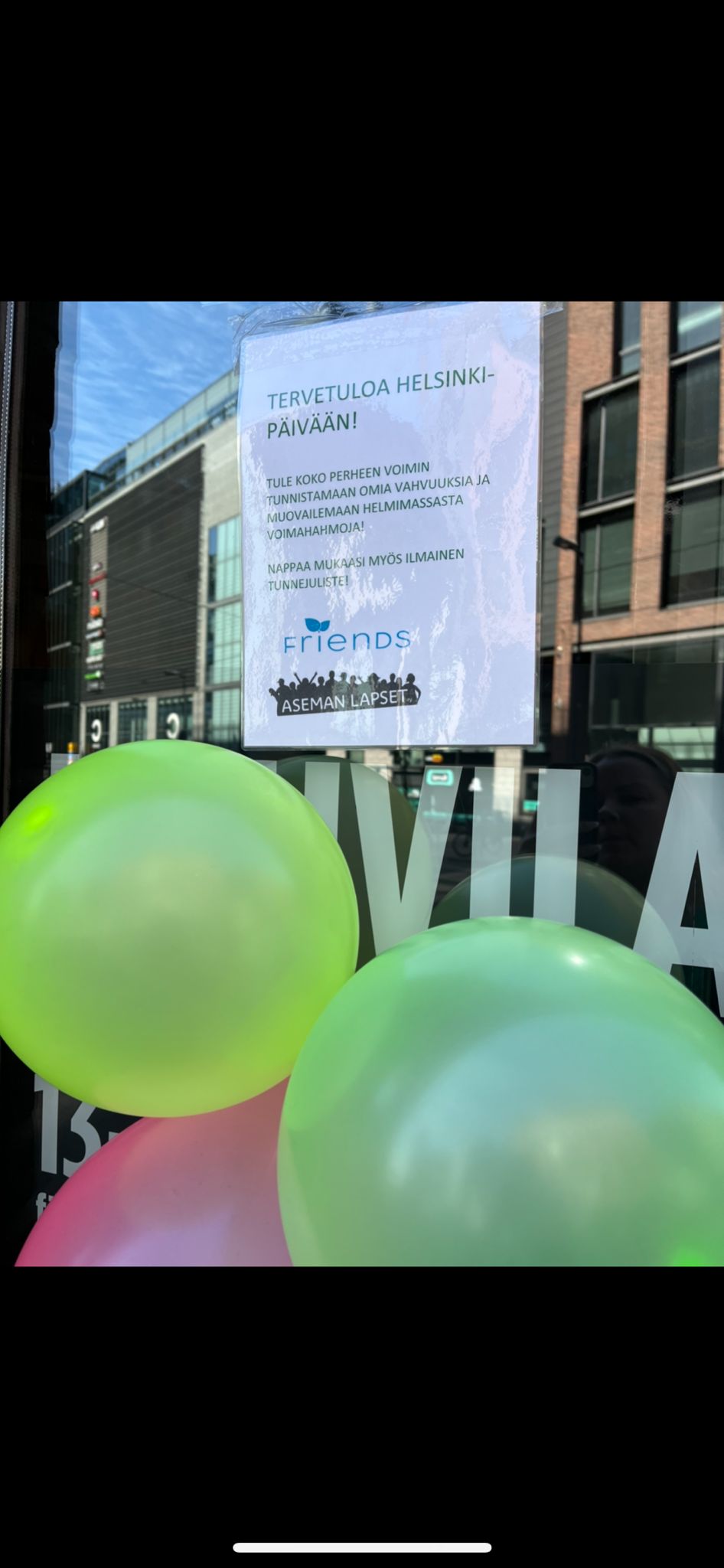 Helsinki-päivän mainos kiinnitettynä ikkunalasiin, edessä värikkäitä ilmapalloja.
