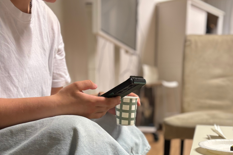 Nuori istuu sohvalla selaten puhelinta, kasvojen yläosa rajautuu kuvasta. Yllään hänellä on valkoinen t-paita ja vaaleat rennot shortsit. Nuorella on nenäkoru ja kaulassa ketju.